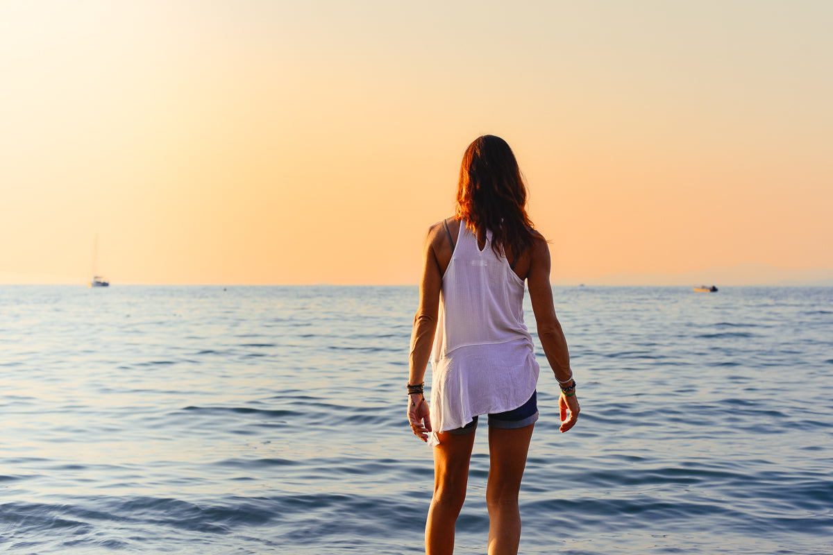woman in denim shorts and flow white top with auburn hair facing the horizon with the sunset and a few boats on the water