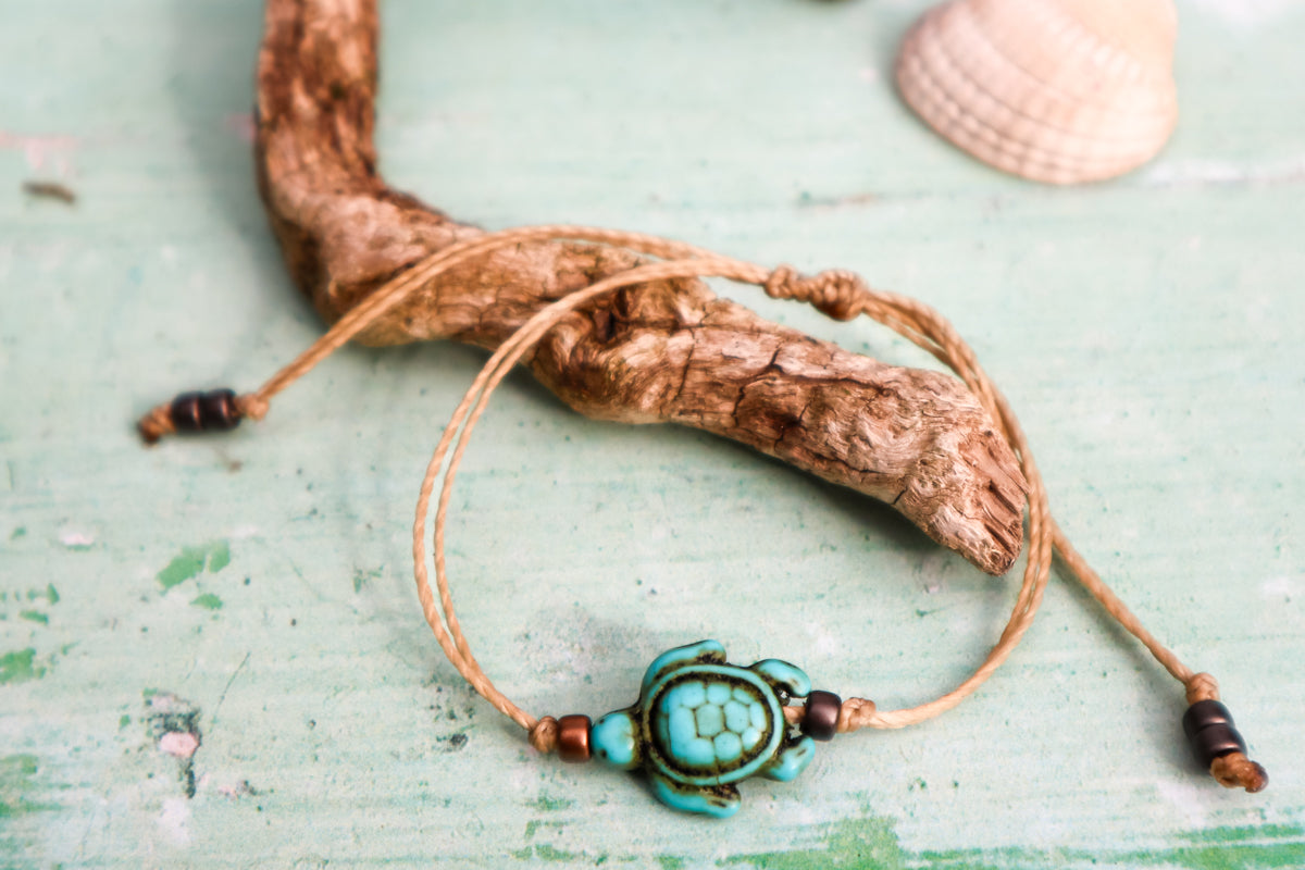 handmade hoplite turtle charm bracelet in blue on a cord with beads styled on a piece of driftwood with a blurred white shell in the background