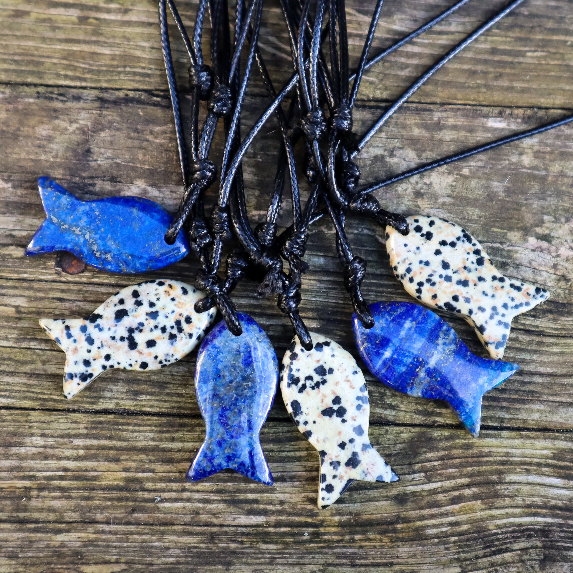 Fish-shaped lapis lazuli and Dalmatian jasper pendants on black cords against a wooden background