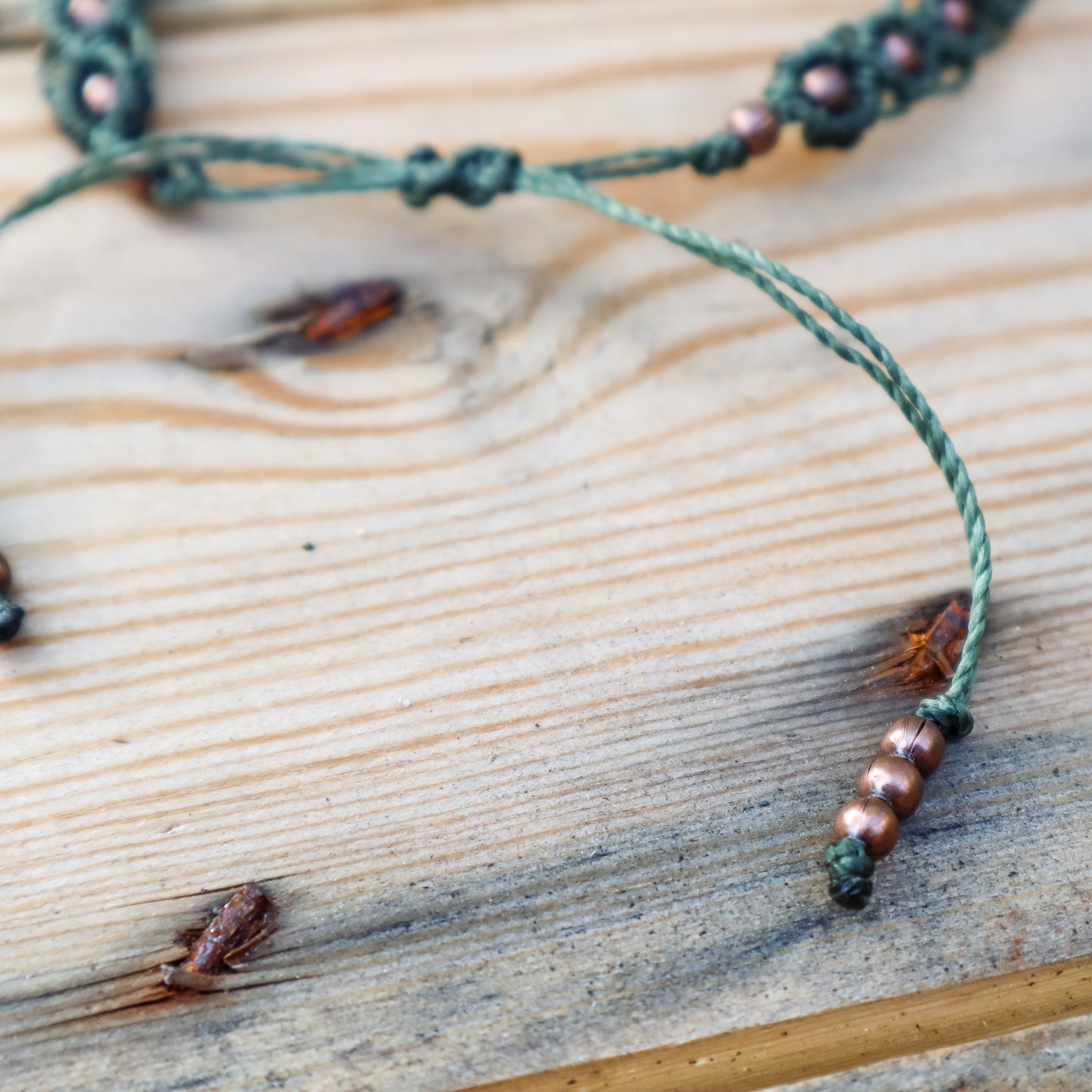Green macrame ankle  bracelet with beads on a wooden surface. Detail of the closure and end cord beads.
