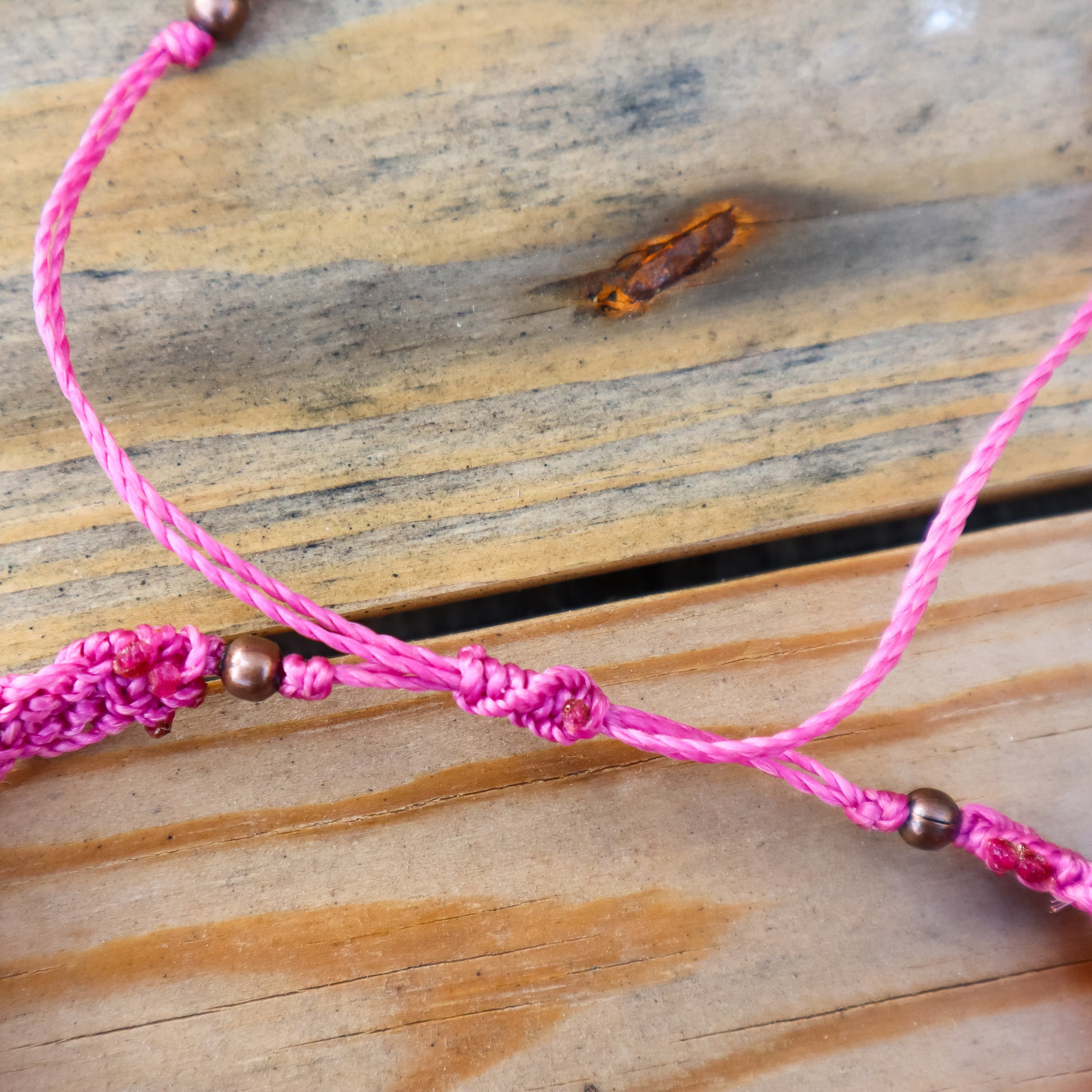 Pink macrame ankle bracelet with bronze beads on a wooden surface. Close up of the sliding knot closure.