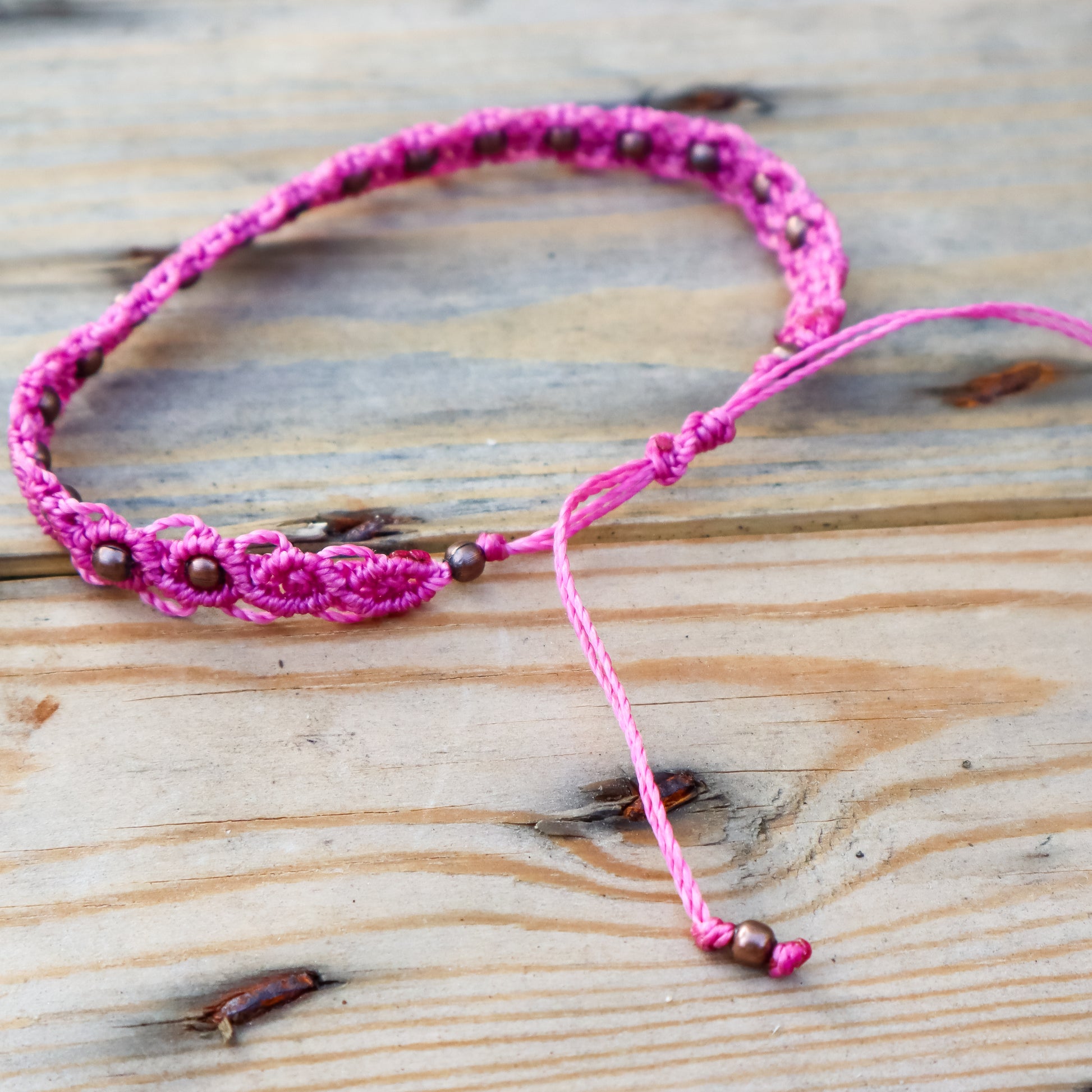 Pink macrame ankle bracelet with bronze beads on a wooden surface