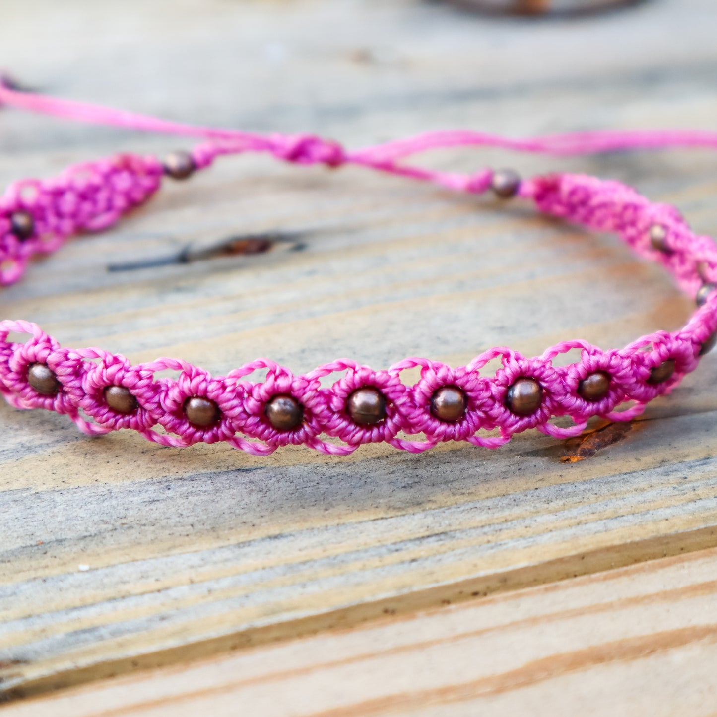 Close up of a pink macrame ankle bracelet with bronze beads on a wooden surface