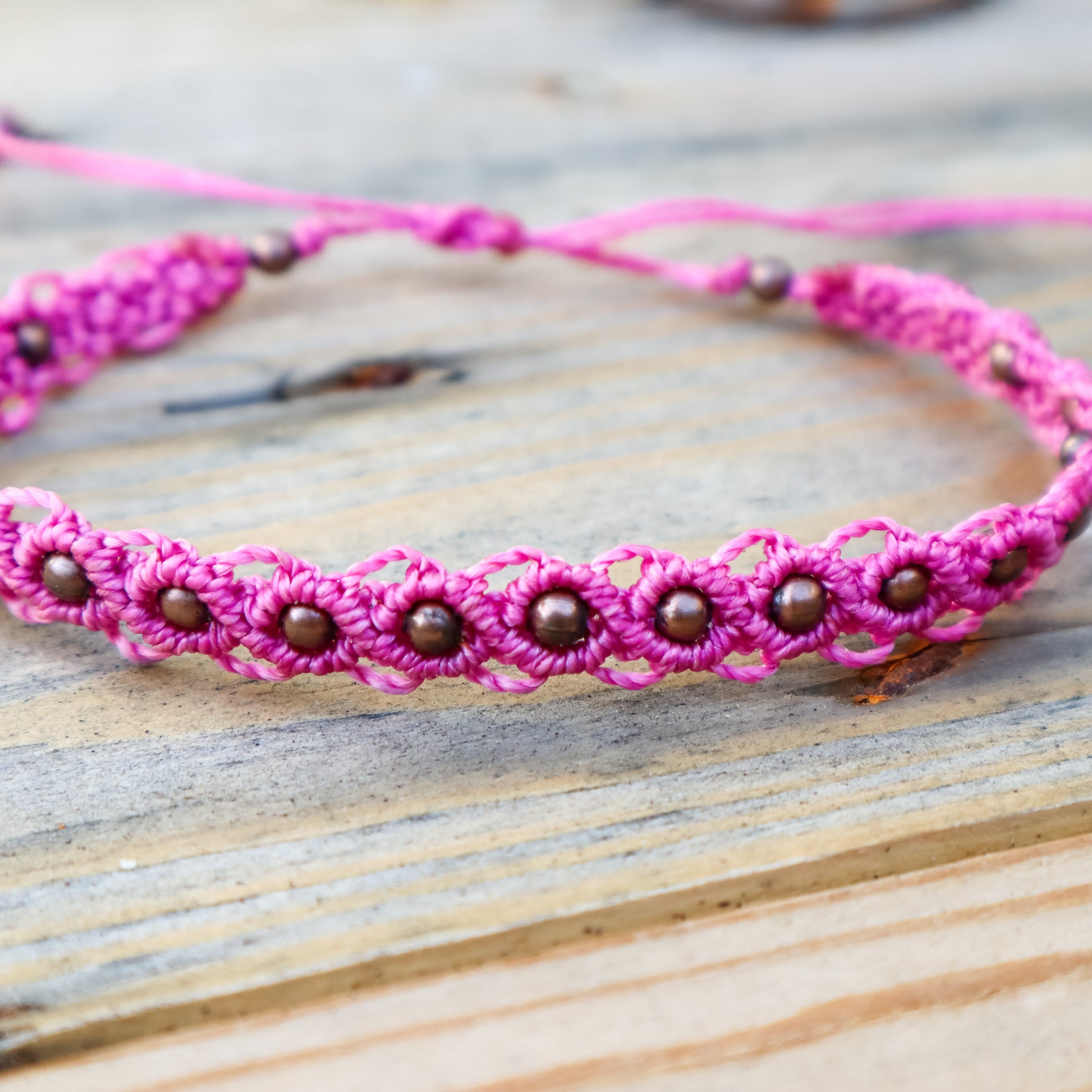 Close up of a pink macrame ankle bracelet with bronze beads on a wooden surface