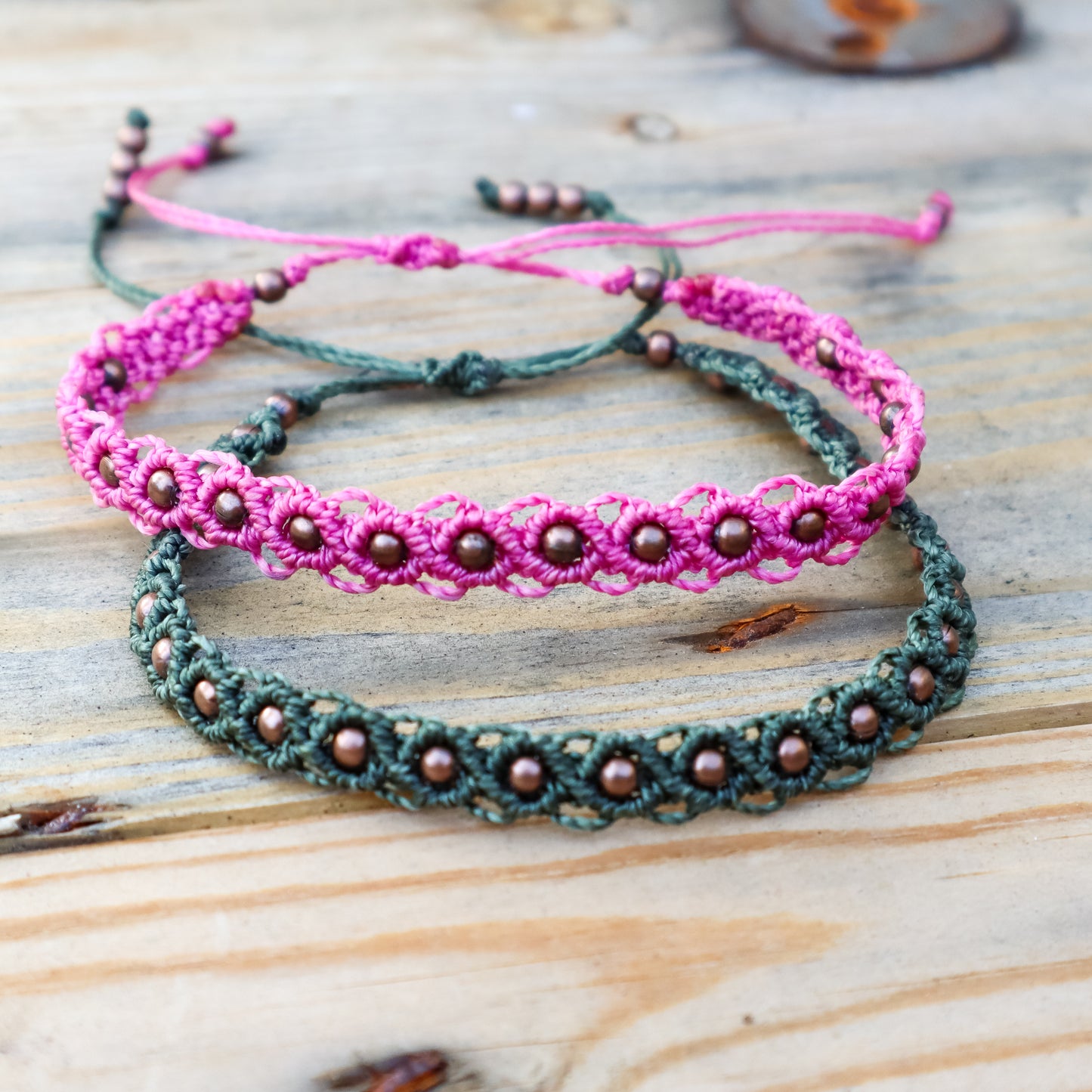 Two  macrame ankle bracelets in green and pink with bronze beads on a wooden surface