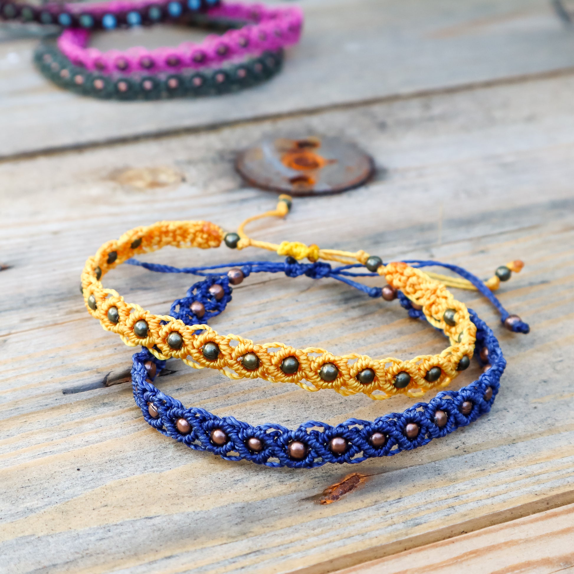 Two macrame ankle bracelets, one blue and one yellow with brown beads, on a wooden surface.