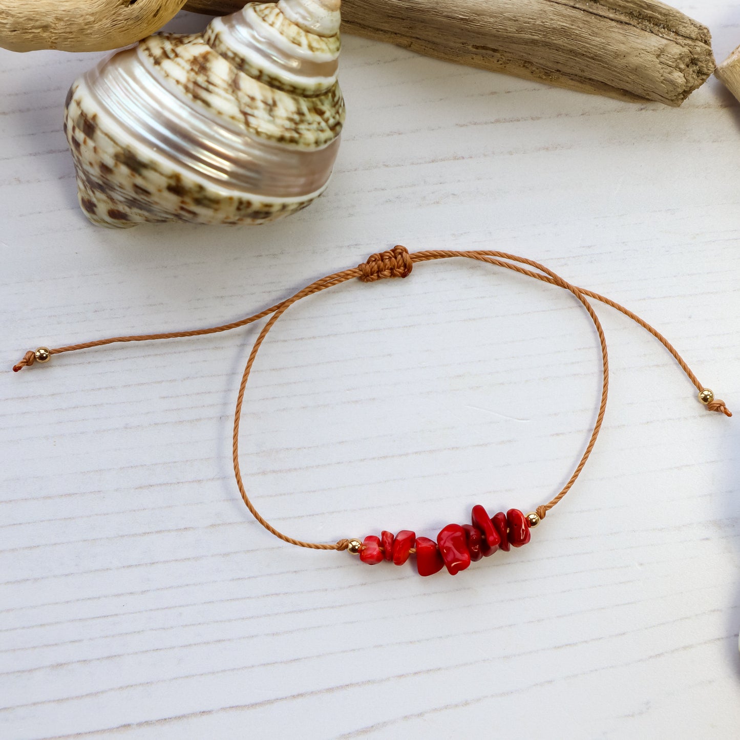 bracelet with red crystal beads on brown cord from above with white wooden background and a shell and driftwood flat lay