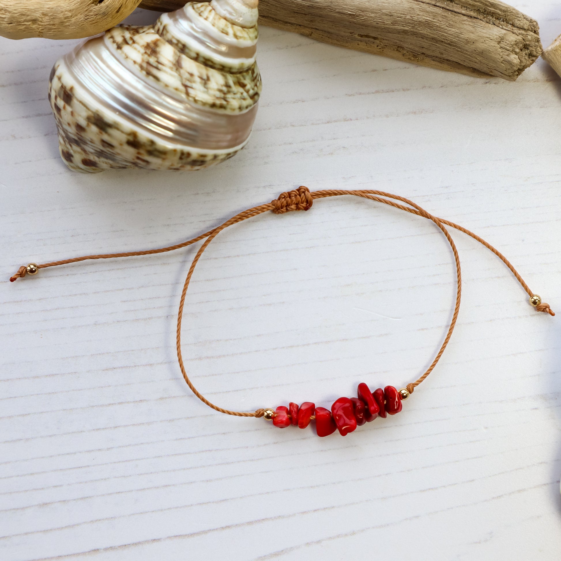 bracelet with red crystal beads on brown cord from above with white wooden background and a shell and driftwood flat lay