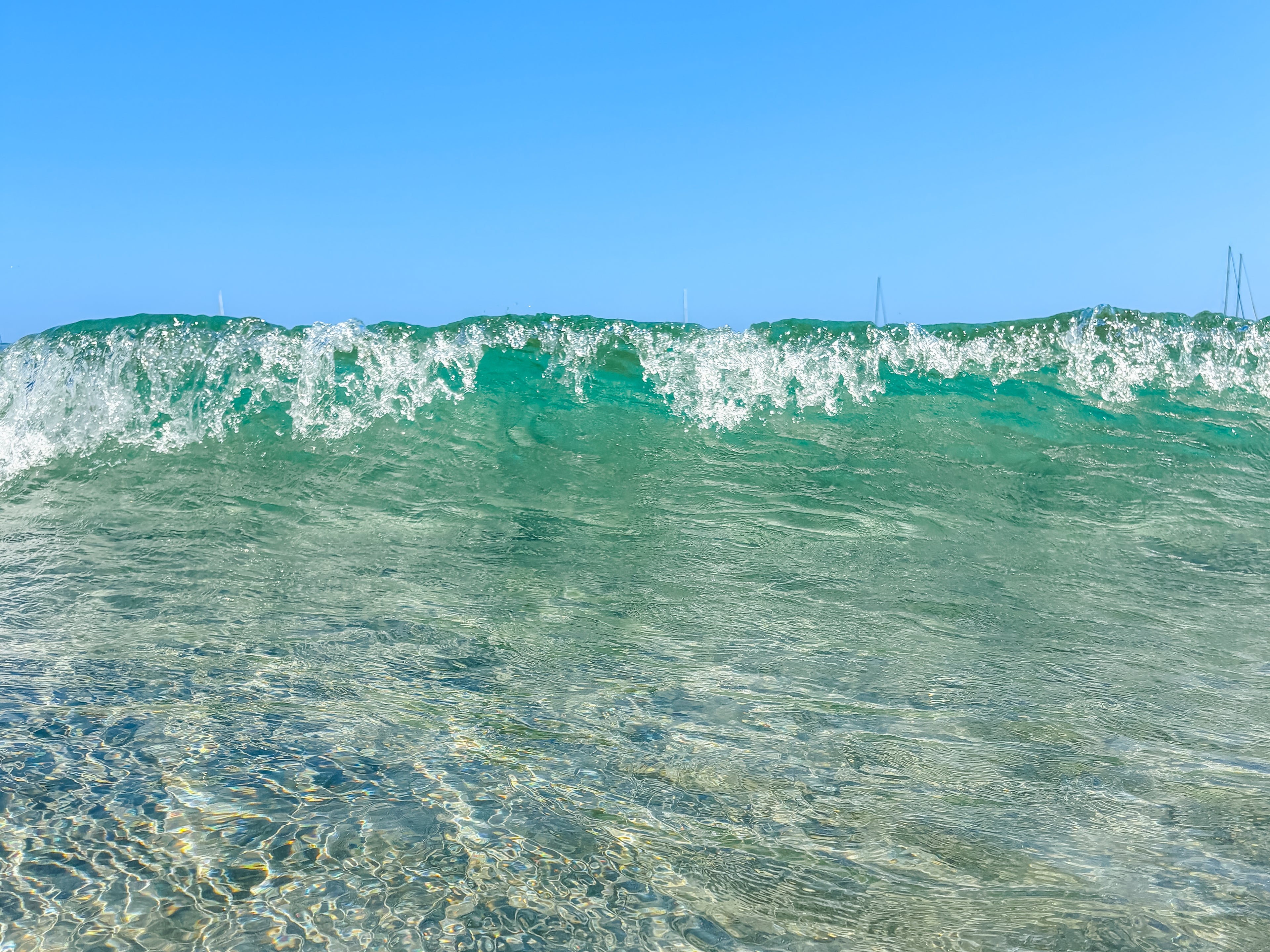 Small wave, turquoise water, sandy bottom, strip of blue sky, sailboats just barely visible beyond the foam of the shallow wave